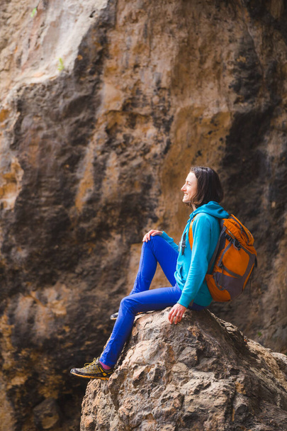 Una mujer con una mochila se sienta en una gran piedra sobre un fondo de montañas rocosas. Viajando solo. La chica subió a la cima y mira a la distancia. - Foto, imagen
