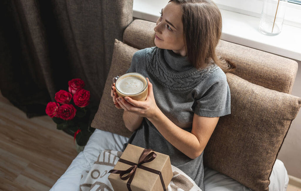 A young beautiful girl is sitting on the bed by the window with a gift box and flowers and enjoying coffee on a festive morning. Concept of a nice start to the day with a cup of cappuccino. - Photo, Image