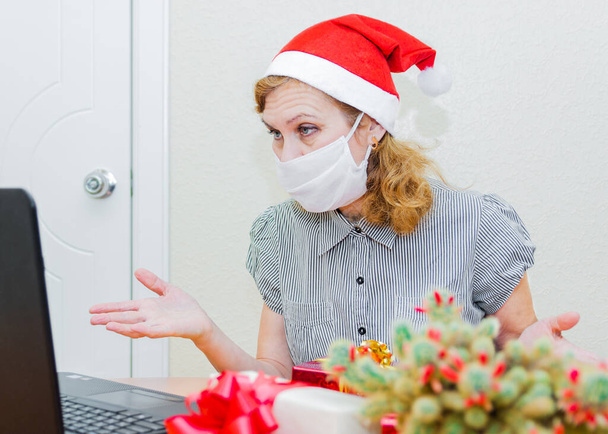 Christmas online greetings. A woman in a medical mask and Santa Claus hat using a computer for video calls congratulates friends and relatives. - Foto, Imagem