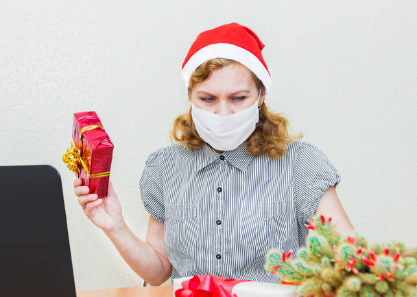 A woman in a medical mask and Santa Claus hat, sitting at a computer, holds a gift in her hand in frustration. Christmas online greetings. - 写真・画像