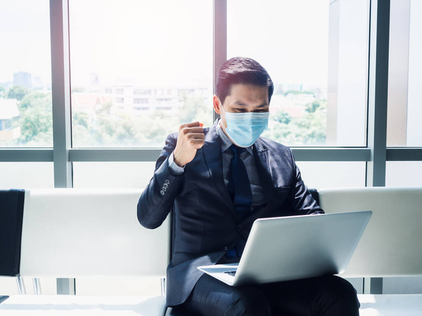 Asiatischer Geschäftsmann im Anzug mit Schutzmaske hob fröhlich die Hand, als er in einem modernen Bürogebäude in der Nähe eines riesigen Glasfensters einen Laptop-Monitor auf seinem Schoß sah.. - Foto, Bild