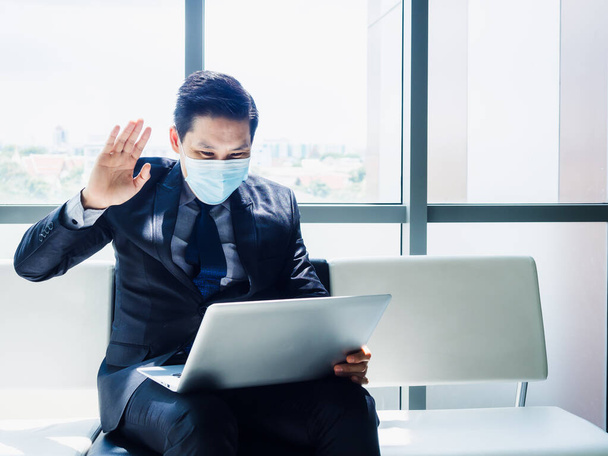 Asiatischer Geschäftsmann im Anzug mit Schutzmaske winkte Kollegen im Laptop-Computermonitor auf seinem Schoß zu, während er in einem modernen Bürogebäude in der Nähe eines riesigen Glasfensters saß, vertikal. - Foto, Bild