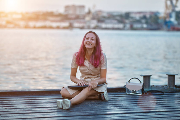 Smiling lady with pink hair sitting on pier with laptop. Lake or river on background. Travel time and new emotion idea - Foto, Imagem