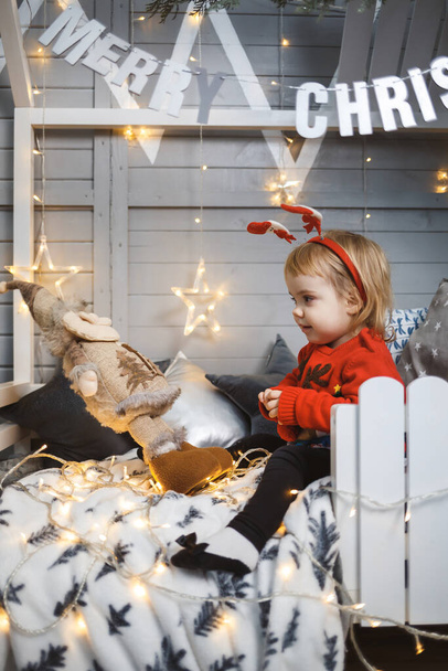 Ein kleines Mädchen im roten warmen Pullover sitzt auf einem Bett neben einem Weihnachtsbaum mit Spielzeug und Geschenken. Glückliche Kindheit. Neujahrsstimmung - Foto, Bild