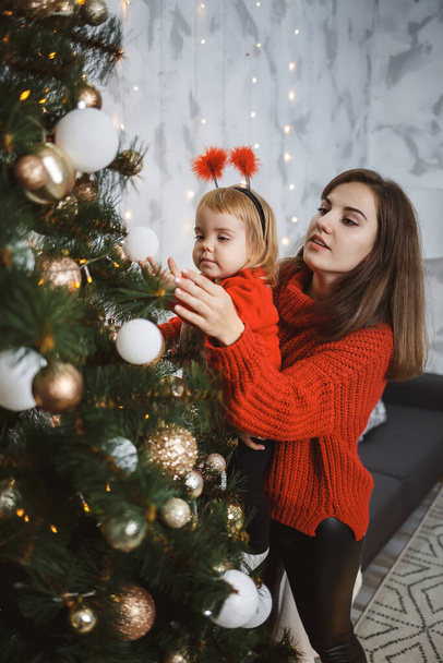 Mom and daughter in red warm sweaters decorate the Christmas tree. Happy motherhood. Warm family relationships. Christmas and New Year's interior. Love. Family concept. - Фото, изображение