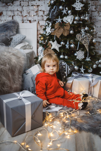 A little girl in a red warm sweater sits under a Christmas tree with toys and gifts. Happy childhood. New Year holiday atmosphere - Foto, imagen