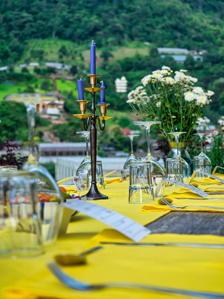 Blue candles on candle holder on the dining table with wine, drinking glasses and yellow placemat. Focus on candles and candle holder. Dinner in nice garden. - Photo, Image