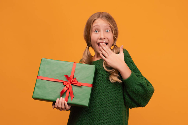 Image of charming blonde girl 12-14 years old in warm green sweater holding present box with red bow. Studio shot, yellow background, isolated. New Year Women's Day birthday holiday concept - Фото, изображение