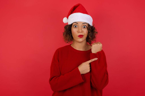 Όμορφη νεαρή Καυκάσια γυναίκα φορώντας χριστουγεννιάτικο καπέλο και κόκκινο πουλόβερ πάνω από το κόκκινο φόντο σε βιασύνη δείχνει να παρακολουθήσουν το χρόνο, ανυπομονησία, αναστατωμένος και θυμωμένος για καθυστέρηση προθεσμία - Φωτογραφία, εικόνα