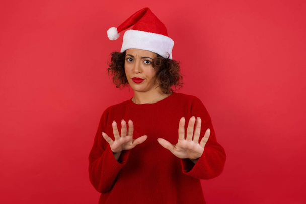 brünette Frau mit Weihnachtsmütze und rotem Pullover vor rotem Hintergrund hat angeekelten Ausdruck Stop-Geste mit beiden Händen - Foto, Bild