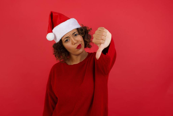 Discontent European woman wearing Christmas hat and red sweater shows disapproval sign, keeps thumb down, expresses dislike, frowns face in discontent, dressed in white shirt, over red background. - Photo, Image
