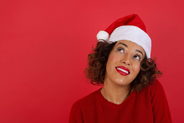 Retrato de una misteriosa mujer con sombrero de Navidad y suéter rojo con el pelo rizado mirando hacia arriba con enigmática sonrisa de pie contra la pared roja - Foto, Imagen