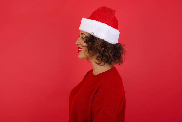 Profiel van lachende vrouw met kerstmuts en rode trui met gezonde zuivere huid heeft contemplatieve expressie, klaar om buiten te lopen over rode studio muur met kopieerruimte - Foto, afbeelding