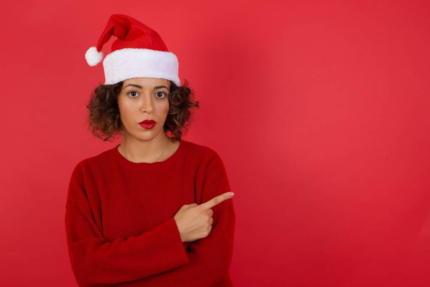 Podekscytowana zaskoczona modna kobieta w kapeluszu świątecznym i czerwonym swetrze wskazuje szczęśliwie na przestrzeni kopii czerwonego tła dla Twojej mody informacji reklamowej odzieży lub tekstu promocyjnego. - Zdjęcie, obraz