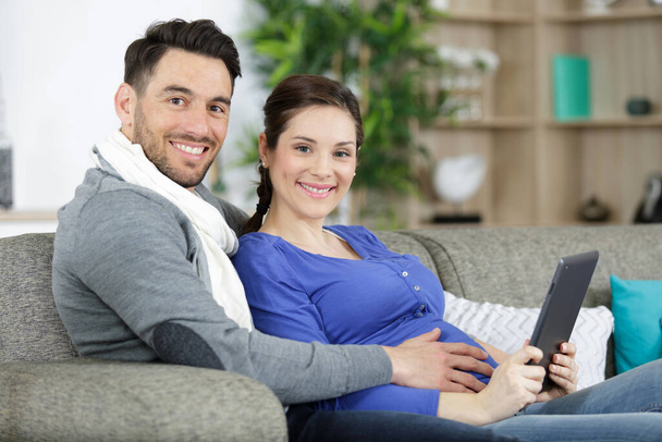 expectant ζευγάρι κάθισε στον καναπέ κοιτάζοντας το tablet PC - Φωτογραφία, εικόνα
