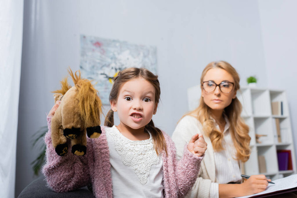 Wütendes kleines Mädchen mit Spielzeug und geballter Faust besucht Psychologin - Foto, Bild