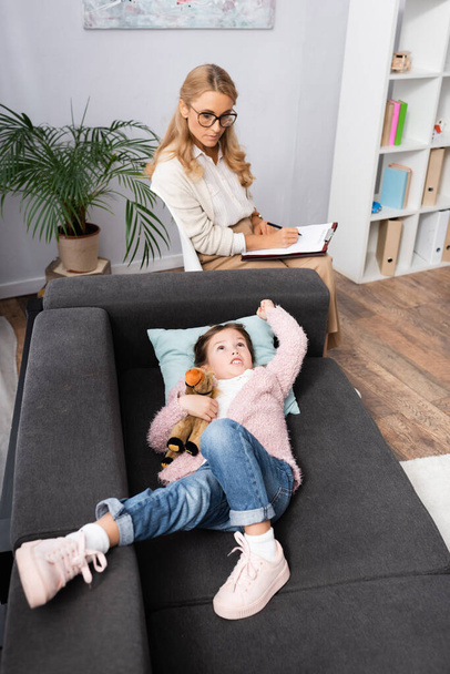 маленькая девочка лежит на диване с игрушкой, рассказывая проблемы психологу - Фото, изображение