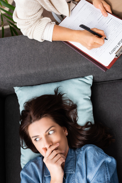 terrified brunette woman covering mouth while therapist writing in medical history - Photo, Image
