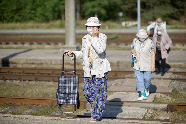 un grupo de viajeros de edad avanzada con máscaras en sus rostros cruzan las vías del tren - Foto, Imagen