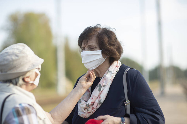 one senior woman helps another put on a medical face mask during a pandemic - Photo, Image