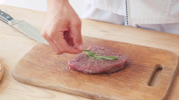 Vista ritagliata dello chef condimento bistecca di manzo con ramo di rosmarino sulla tavola - Filmati, video