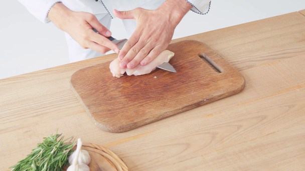 Vista ritagliata dello chef che taglia il filetto di pollo vicino agli ingredienti sul tavolo su bianco - Filmati, video