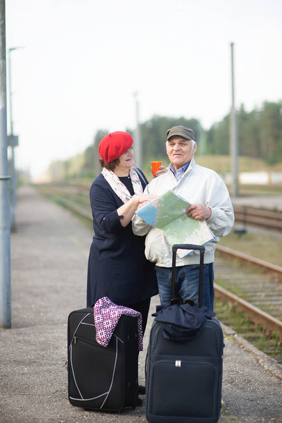 地図を見て電車が旅に出るのを待っているスーツケースを持った幸せな先輩夫婦 - 写真・画像