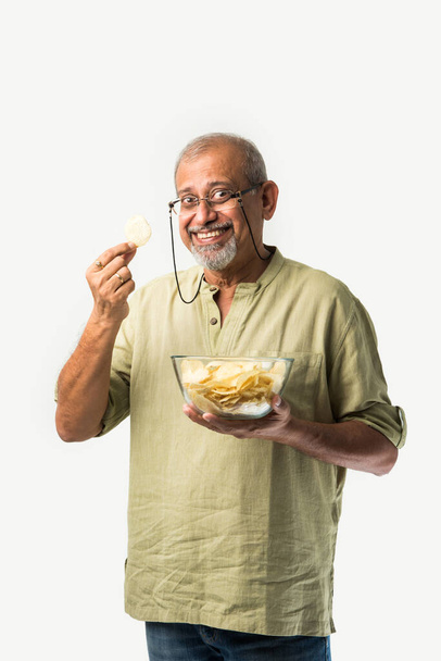 Indian asian old or retired senior man eating potato chips on white background - Photo, Image