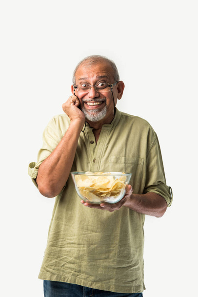Indiaas aziatisch oud of gepensioneerd senior man eten aardappel chips op witte achtergrond - Foto, afbeelding