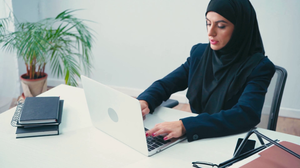 Мусульманская предпринимательница использует ноутбук рядом с бумагами и ноутбуками в офисе  - Кадры, видео