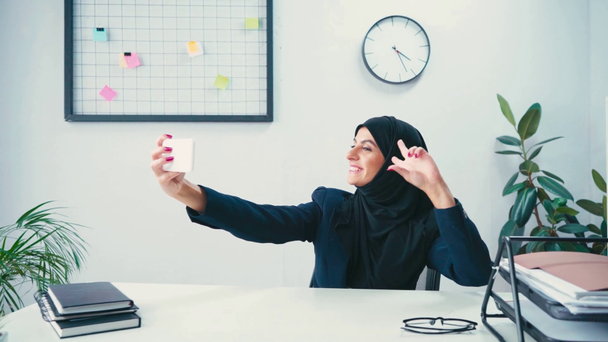Sorridente donna d'affari musulmana che fa gesti mentre si fa selfie su smartphone  - Filmati, video