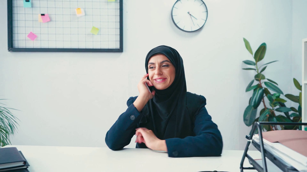 Usmívající se muslimská obchodnice mluví na smartphone v blízkosti papíry v kanceláři  - Záběry, video