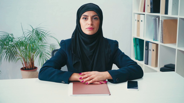 Donna d'affari musulmana guardando la fotocamera vicino alla cartella di carta e smartphone  - Filmati, video