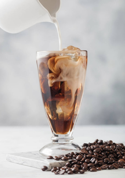 Pouring milk into milkshake classic glass with iced black coffee and fresh raw beans on light table background. - Zdjęcie, obraz