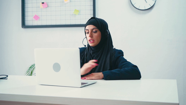 Donna d'affari musulmana in auricolare utilizzando il computer portatile in ufficio  - Filmati, video
