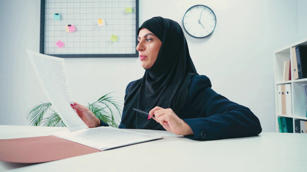 Femme d'affaires musulmane souriante avec stylo et papiers parlant à la caméra dans le bureau  - Séquence, vidéo