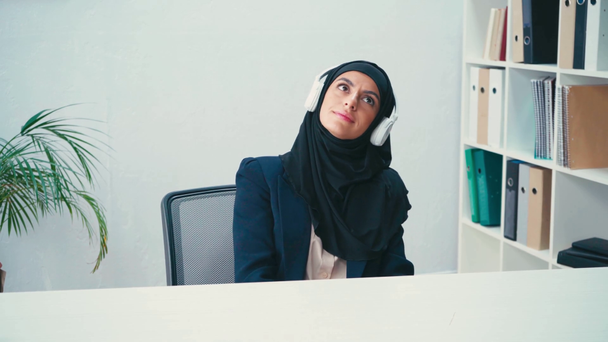 Femme d'affaires musulmane dans le hijab et écouteurs assis dans le bureau  - Séquence, vidéo