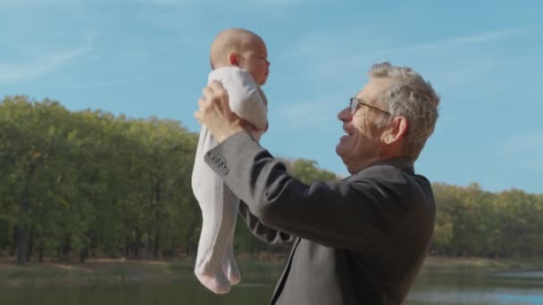 Anciano mayor hablando con su nieto bebé, sosteniéndolo en el aire, de cerca, vista lateral. - Metraje, vídeo