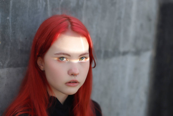 Genç bir kızın portresi piercingli ve kızıl saçlı gri beton bir duvara yaslanmış, gözlerinde güneşli bir ışınla - Fotoğraf, Görsel