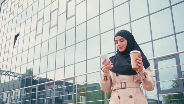 Usmívající se muslimka pomocí smartphone a pití kávy jít blízko budovy  - Záběry, video