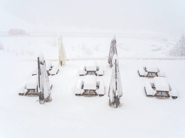 Snow-covered tables with benches and folded outdoor umbrellas stand on the site on a foggy snowy day - Photo, Image