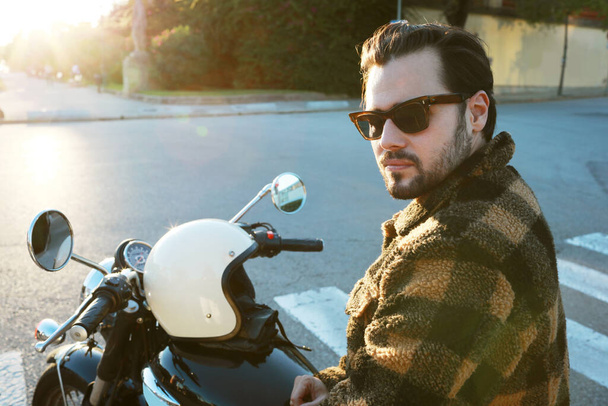 Portrait of young bearded biker man on motor bike posing outdoors at the city streets. - Photo, image