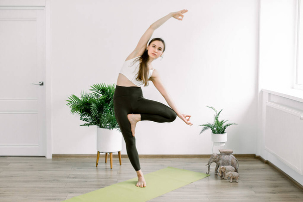 Young attractive Caucasian woman practicing yoga, standing in Vrksasana exercise, Tree pose with mudra, working out in modern light fit hall. Indoor horizontal studio image - Φωτογραφία, εικόνα