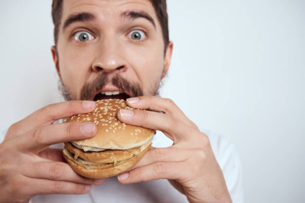 A man eating a hamburger on a light background in a white T-shirt cropped view close-up hunger fast food - Foto, afbeelding