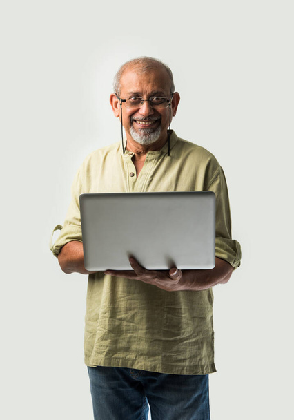 Indian asian senior adult man or grandpa using laptop and holding paper currency notes against white background - Foto, afbeelding