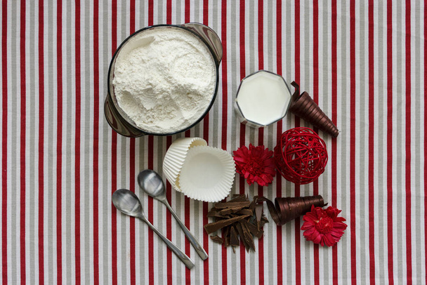 Zutaten zum Backen von Cupcakes - Mehl, Milch, Schokolade. Weiße Papierformen auf der Tischdecke mit grauen, roten und weißen Streifen. Vorbereitung auf das Backen. Ansicht von oben. - Foto, Bild