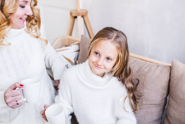 Mother and daughter, girl sitting on the sofa in light sweaters and drinking tea from a mug, bright interior, winter atmosphere, on the eve of Christmas - Foto, Bild
