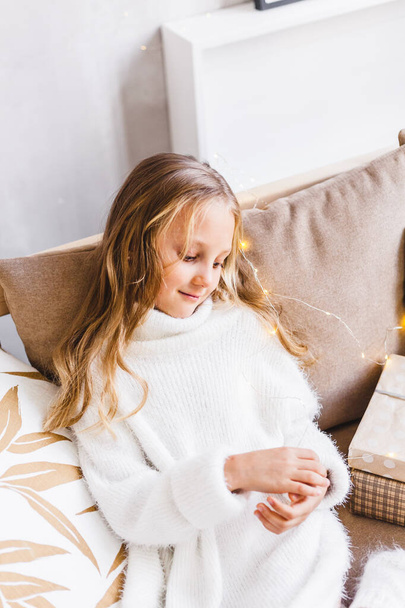 Girl, daughter, baby sitting on the sofa in a light warm sweater, long blonde hair, European appearance and light interior - Photo, Image