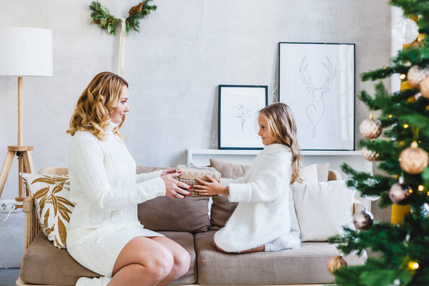 Mom daughter blonde hair dressed in light sweaters, waiting holiday, room decorated celebrate Christmas, sitting couch, mom gives gift daughter - Foto, immagini