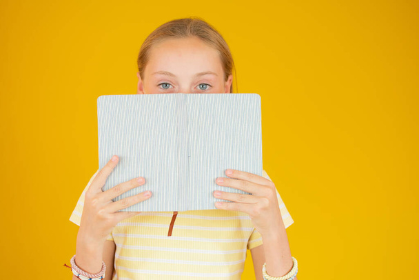 Foto van kind meisje verbergen haar lippen met boek geïsoleerd over gele achtergrond - Foto, afbeelding